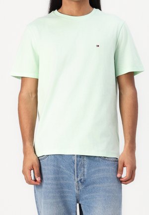 Mężczyzna ubrany w jednolity, jasnozielony t-shirt z krótkim rękawem i niebieskie dżinsy, stoi z rękami swobodnie opuszczonymi wzdłuż ciała.
