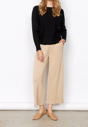 Schwarzes Langarmoberteil, beiger weit geschnittener Hosen und brauner Slipper. Die Stoffe sind glatt und leicht. Die Hose hat einen elastischen Bund und eine lockere Passform.
