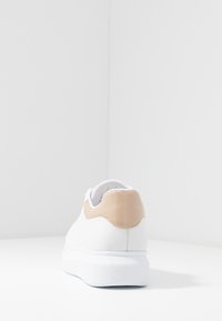 Bílá teniska s hladkým koženým svrškem, béžovým akcentem na patě a texturovanou bílou podrážkou. Jednoduchý a minimalistický design.