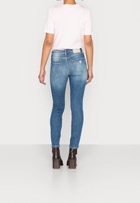 Jeans de mezclilla azul ajustados con un lavado desgastado, que cuentan con dos bolsillos traseros y un pequeño detalle del logo en la cintura. Combinados con una camiseta rosa y botas de tacón marrones.
