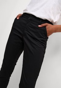 Pantalon noir à coupe slim, fabriqué en tissu doux, avec des poches latérales et une texture lisse. Conçu avec une taille mi-haute.