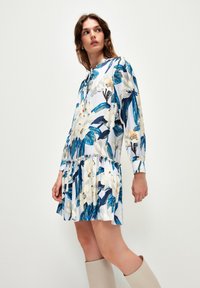 Blumenkleid in Weiß, Blau und Grün. Mit langen Ärmeln, geraffter Taille und gemustertem Stoff mit Blättern und Blüten.