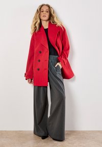 Punainen kaksoisnapitettu takki suurilla mustilla napituksilla, yhdistetty leveälahkeisiin harmaisiin raidoitettuihin housuihin. Sujuva tekstuuri, nykyaikainen muotoilu.