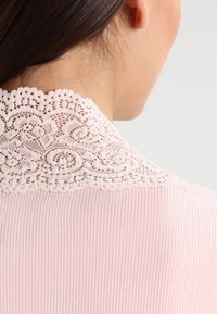 Ljust rosa blus i ett texturerat ribbat tyg med en detaljerad spetskrage. Kragen är i cremefärg med intrikata mönster.