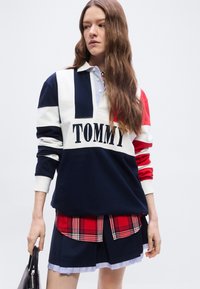 Νεαρή γυναίκα με μακριά κυματιστά μαλλιά φοράει μπλε, λευκό και κόκκινο φούτερ με το γράμμα "TOMMY" πάνω από κόκκινο καρό πουκάμισο και σκούρο μπλε πλισέ φούστα, κρατώντας μια μαύρη τσάντα.