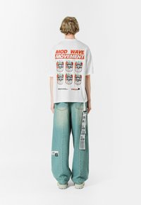 Camiseta blanca de gran tamaño con un estampado gráfico que presenta seis diseños de latas y el texto "MOVIMIENTO OLA MOD". Pantalones de mezclilla holgados en azul claro con etiquetas.