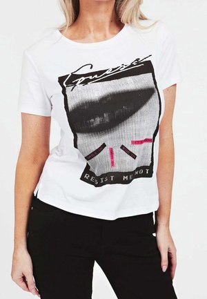 Femme portant un t-shirt blanc avec un graphisme noir de lèvres et du texte incluant « Resist Me Not » ainsi que de petites étiquettes roses sur le devant.