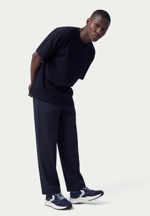 Man leaning forward wearing a loose black t-shirt, pinstriped navy pants, and navy-white sneakers against a plain background.