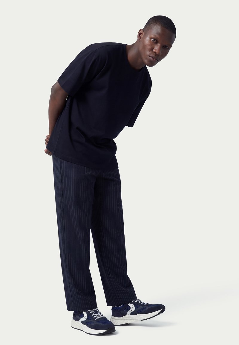 Mann lehnt nach vorne, trägt ein lockeres schwarzes T-Shirt, marineblaue Hosen mit Nadelstreifen und marineblaue Sneakers mit weißen Details vor einem schlichten Hintergrund.