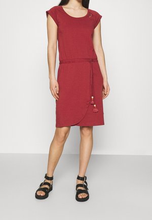 Frau trägt ein rotes, knielanges, kurzärmeliges Kleid mit Kordelzug in der Taille und schwarze Sandalen mit Knöchelriemen, steht vor einem schlichten Hintergrund.