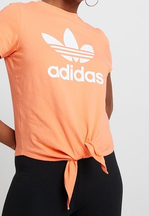 Osoba ubrana w brzoskwiniową koszulkę Adidas zawiązaną w talii oraz czarne spodnie, stojąca na jednolitym jasnym tle.