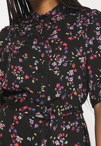 Černé květinové šaty s límečkem, krátkými rukávy a zavazovacím pasem. Látka zobrazuje barevné květy v růžové, modré a žluté barvě.