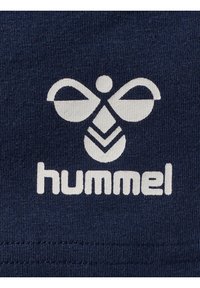 Granatowy materiał z białym logo Hummel, które zawiera stylizowane skrzydła oraz geometryczny wzór. Gładka tekstura z detalami przeszycia.