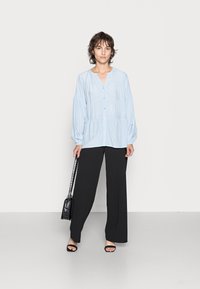 Camisa azul-claro de mangas compridas com decote em V e detalhes plissados, combinada com calças largas pretas e sandálias pretas de tiras. Bolsa preta transversal.