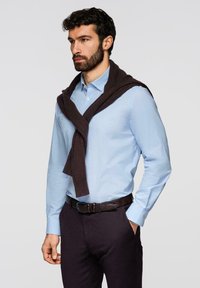 Homme barbu portant une chemise habillée bleu clair et un pantalon foncé avec un pull sombre décontracté posé sur les épaules, debout avec une main dans la poche.