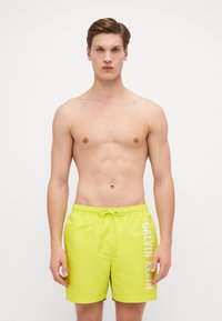 Shorts de bain jaunes vif en matériau léger, dotés d'une taille élastique avec cordon de serrage et d'un imprimé logo blanc sur le côté.