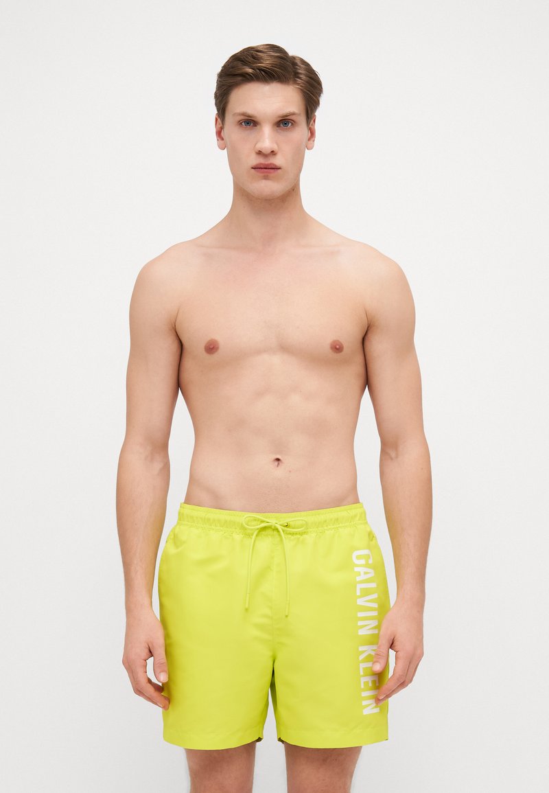 Shorts de bain jaunes vif en matériau léger, dotés d'une taille élastique avec cordon de serrage et d'un imprimé logo blanc sur le côté.