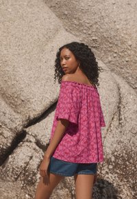 Lilla off-the-shoulder top med blomsterprint, flagrende ærmer, elastisk halsudskæring, kombineret med denimshorts foran en klippefyldt baggrund.