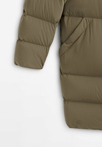 Manche et côté d'une veste d'hiver matelassée vert olive avec poche à couture diagonale visible, sur fond blanc.