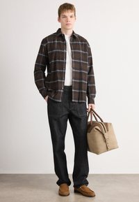 Junger Mann in kariertem braunen Hemd, weißem T-Shirt, schwarzen Hosen und hellbraunen Slippern, der eine beige und braune Reisetasche hält und vor einem schlichten weißen Hintergrund steht.
