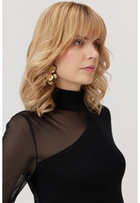Czarny top o wysokim dekolcie, z jedną przezroczystą rękawą i teksturowanym materiałem. Modelka nosi złote kwiatowe kolczyki, co stanowi kontrastowy detal.