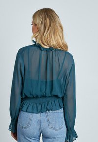 Blusa trasparente turchese con maniche lunghe, scollatura arricciata e vita elasticizzata. Indossata su jeans in denim blu con tasche posteriori visibili.