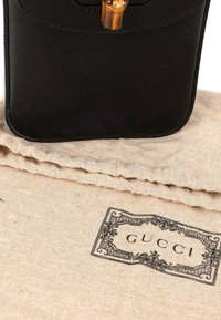 Sac à main en cuir noir avec fermoir doré sur pochette en tissu beige imprimée du logo Gucci et bordure décorative.