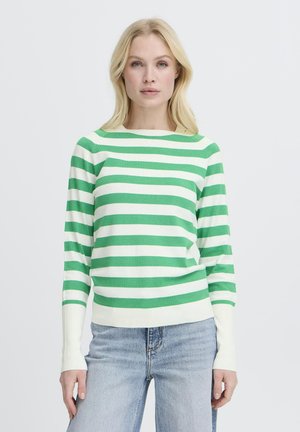 Mujer rubia con un suéter de rayas verdes y blancas de manga larga y jeans azul claro contra un fondo blanco liso.