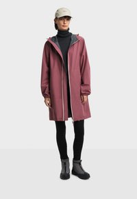 Žena nosi rađeni kaput u prašnjavoj ružičastoj boji preko crnog dukserice i tajica, sive gležnjače i bež kapu s logotipom.