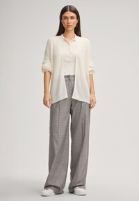 Donna in piedi che indossa un cardigan bianco lavorato a maglia sopra una polo crema, pantaloni a quadri grigi a gamba larga e sneakers bianche su uno sfondo neutro.