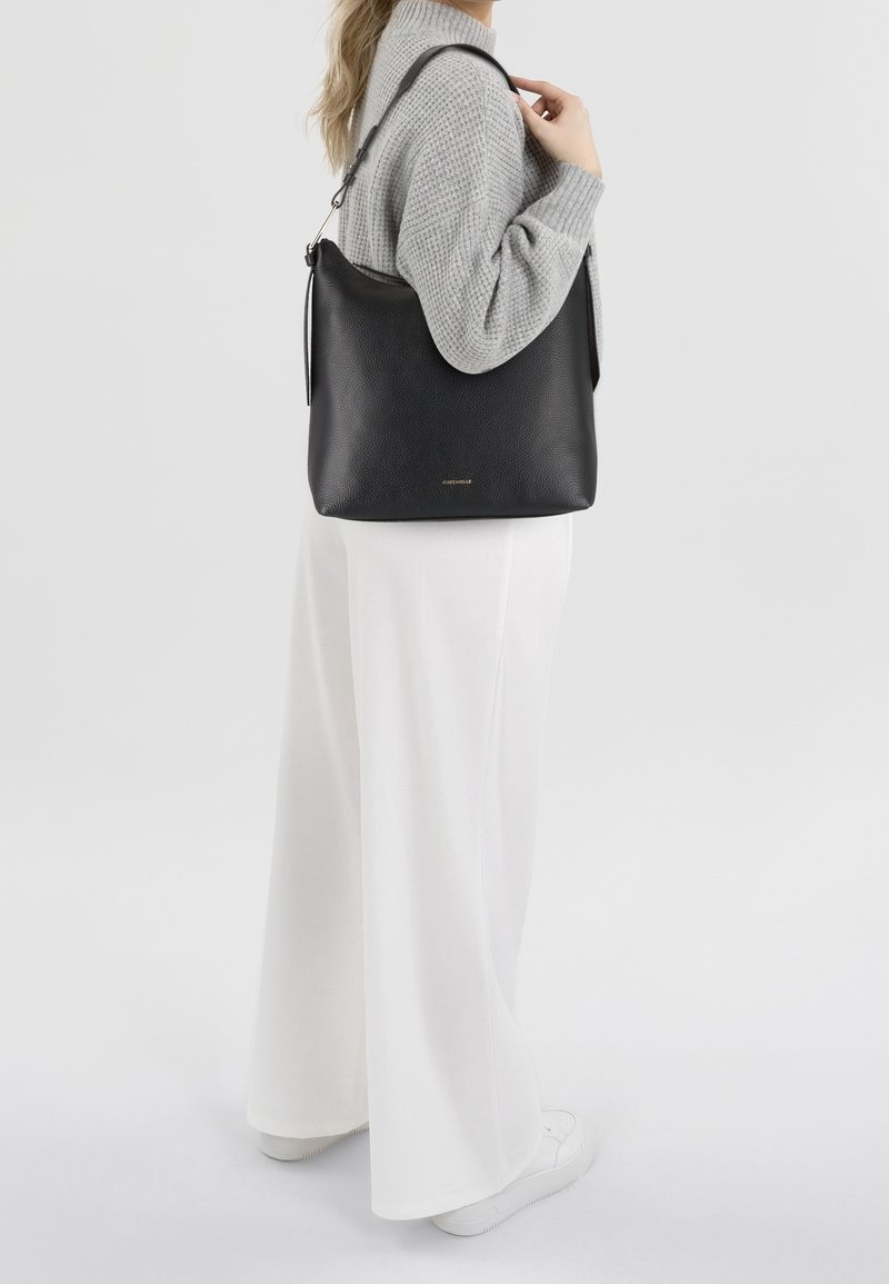 Femme en pull gris et pantalon blanc large tenant un sac à bandoulière en cuir noir avec logo doré sur fond uni.