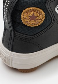Zapatilla negra con parte superior de cuero texturizado y tela, que presenta un parche marrón circular con un logo de estrella, y suela de goma blanca con agarre.