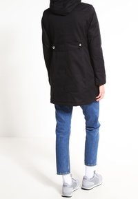 Svart ytterjacka med huva, med insvängd midja, tryckknappar och en slät, matt yta. I kombination med blå denim och grå sneakers.