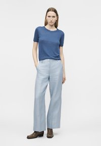 Femme debout, les mains dans un pantalon large bleu pâle, portant un haut en maille bleu à manches courtes et des bottines marron sur fond blanc.