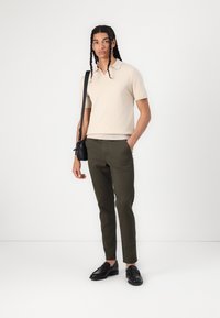 Polo shirt en tricot beige à manches courtes, pantalons verts olive et mocassins noirs. Le modèle tient un sac banane noir. Design simple.
