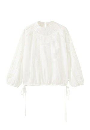 Weiße Bluse mit langen Ärmeln, Spitzenverzierung am Einsatz und an den Ärmeln, Kellerfalten vorne, elastischem Saum mit seitlichen Bändern und Knopfverschluss mit Schlitz am Nacken.