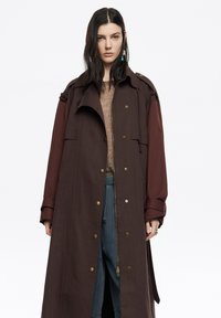 Długi, dwukolorowy płaszcz trencz w ciemnym brązie i bordowym, z dużymi guzikami, kołnierzem i bocznymi kieszeniami. Materiał wydaje się teksturowany i wytrzymały.