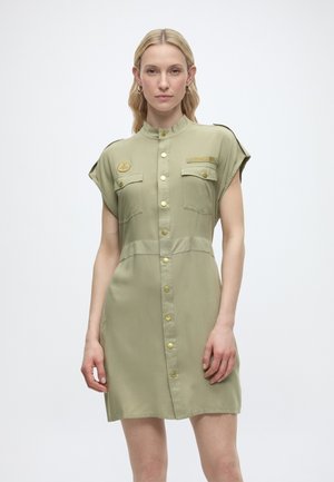 Frau mit blonden Haaren, die ein kurzärmliges, khakifarbenes Kleid mit Brusttaschen und goldenen Knöpfen trägt, steht vor einem einfarbigen Hintergrund.