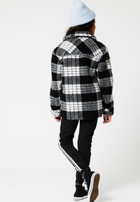 Chaqueta de forro polar a cuadros en blanco y negro con un corte recto, que presenta un cuello, mangas largas y una superficie texturizada, combinada con pantalones negros y zapatillas.