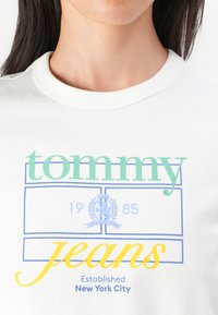 T-shirt en coton blanc avec des graphiques colorés : "tommy" en vert, contours bleus, "jeans" en jaune, insigne 1985, et "Établi à New York City."