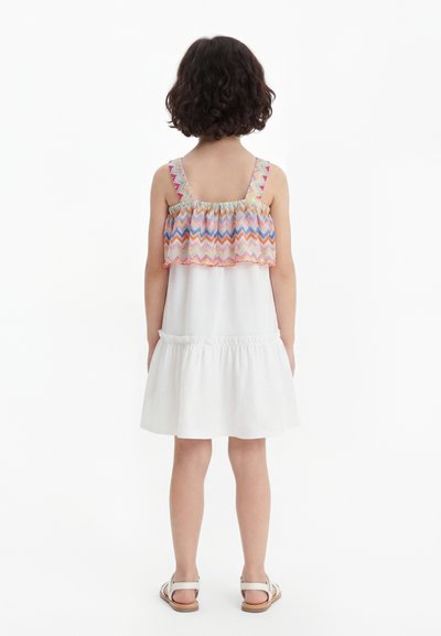 Παιδί με κοντά σγουρά μαλλιά που φοράει λευκό αμάνικο φόρεμα με πολύχρωμο ζιγκ ζαγκ βολάν και λευκά σανδάλια, στέκεται με την πλάτη στραμμένη.