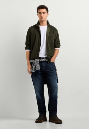 Jeune homme debout, les mains dans les poches, portant une chemise vert foncé sur un t-shirt blanc, un jean bleu, un pull rayé noué à la taille et des bottes foncées.