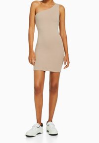 Kvinna klädd i en beige, enaxlad bodycon-miniklänning och vita sneakers med svarta sulor, som står mot en enfärgad vit bakgrund.