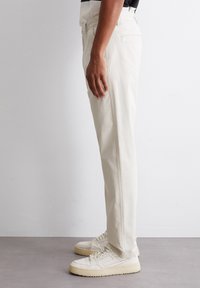 Hellebeige Baumwollhose mit geradem Bein, seitlichen Taschen und einer glatten Textur. Getragen mit weißen Sneakers.