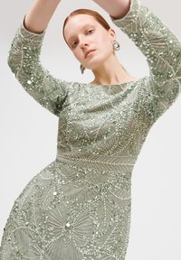 Grünes Perlenkleid mit einer taillierten Silhouette, langen Ärmeln und aufwendigen Blattmustern aus Pailletten und Perlen auf transparentem Stoff.