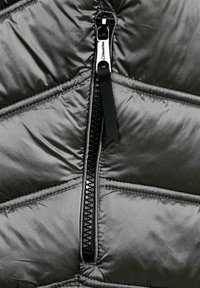 Grå puffjacka med en svart dragkedja, quiltad textur och glänsande material. Dragkedjans dragflik är i tyg.