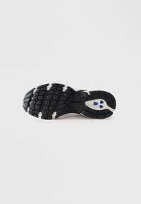 Semelle de chaussure de sport noire et blanche avec des motifs de grip profonds et deux petites accents bleues près du talon sur un fond blanc.