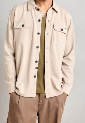Man wearing beige button-up shirt over olive ribbed tank top and brown pants, hands in pockets, with tattoo visible on upper chest.