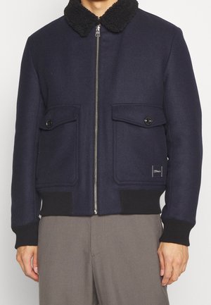 Marineblaue Bomberjacke mit schwarzem Sherpa-Kragen, Frontreißverschluss, zwei Taschen mit Knopflappen und schwarzen gerippten Bündchen und Saum an einer Person mit grauer Hose.