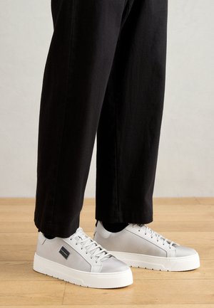 Grijze mesh sneakers met witte rubberen zolen, voorzien van platte veters en een zwart logo-label aan de zijkant, gepaard met zwarte broek.